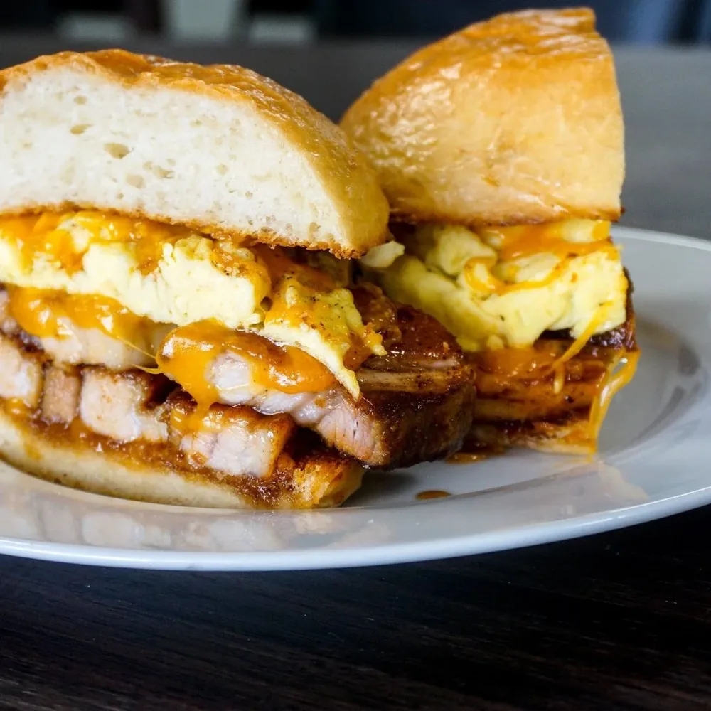 A pork belly, egg and melted cheese sandwich on a toasted roll, cut in half.